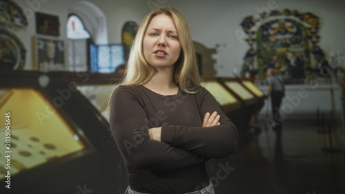 Canvas Print Woman blonde with arms crossed pouting beside glass displaycases in a museum exhibition hall with other visitors nearby; frustration defiance isolation
