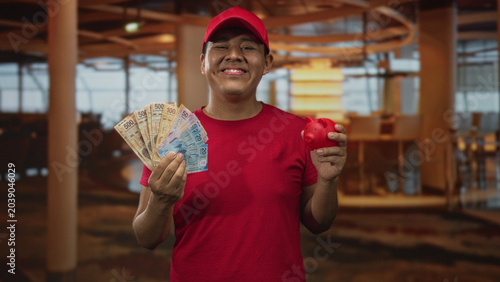 Valokuva Hispanic man holding fan of pesos and red piggybank, smiling and holding money in a restaurant building, wearing red cap and red shirt; financial pride