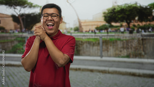 Fotografija Hispanic man clasping hands and laughing on a cobblestone street near a historic building, wearing red polo and glasses; joy celebration