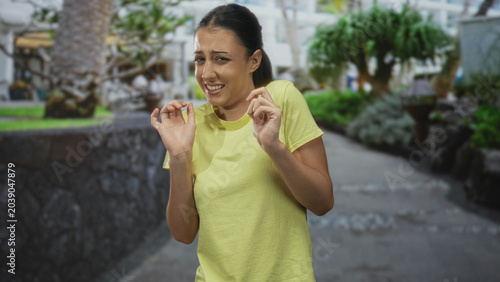 Fotografi Young hispanic woman with hands raised recoiling on a street walkway wearing a yellow t shirt; disgust aversion