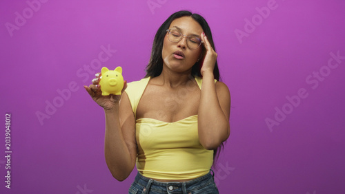 Tablou pe pânză Woman holds yellow piggybank, hand on temple, wearing glasses and yellow top in studio; saving dilemma concern