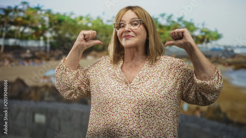 Konstfotografi Woman pointing thumbs to chest and smiling, wearing floral blouse and glasses, standing in proud pose in studio with beach backdrop; confidence empowerment
