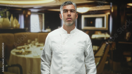 Obraz na plátne Man chef in white jacket presses palms together by a set dining table in building; humble pleading appeal