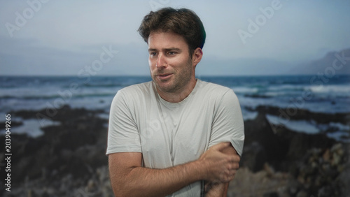 Fotografía Man holds his arm on a rocky seaside coast in a plain white tshirt, grimacing and shifting posture while leaning forward, looking tense; anxiety