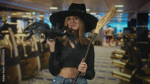 Foto Woman holds faux spider near face while gripping broom and showing midriff in a building dressed as a witch; playful halloween fun