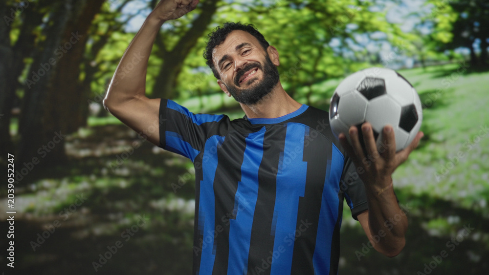 Obraz premium Man bearded soccer player in blue uniform holds soccer ball and flexes bicep while smiling in forest; pride.