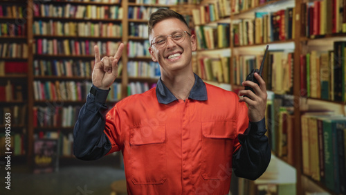 Fotografia Man mechanic in orange coverall holding a walkie talkie and making rock horns gesture with his hand in a building library; playful joy