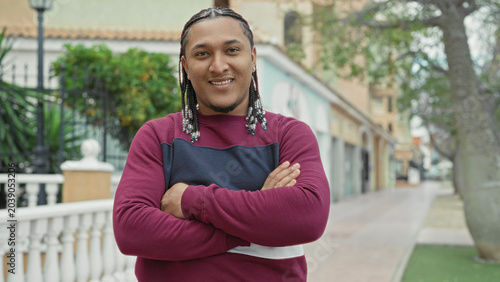 Foto Man with beaded braids smiling with arms crossed on a street by white balustrade and storefronts; confidence approachability