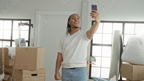 Bilde på lerret Man in white polo holds smartphone aloft taking a selfie among moving boxes inside an apartment building; new home optimism