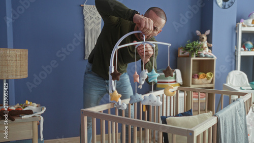Foto Man leaning over wooden cradle adjusting plush mobile above mattress in studio baby room; bonding tenderness