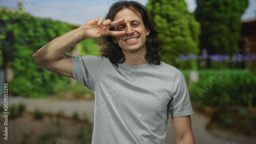 Young hispanic man smiling and holding two fingers in a peace sign over his eye in a green park pathway; playful joy.