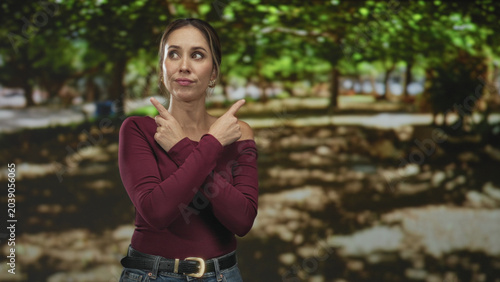 Obraz na plátne Woman points index fingers to opposite sides with crossed arms, wearing burgundy sweater and jeans, standing before trees in forest; indecision choice