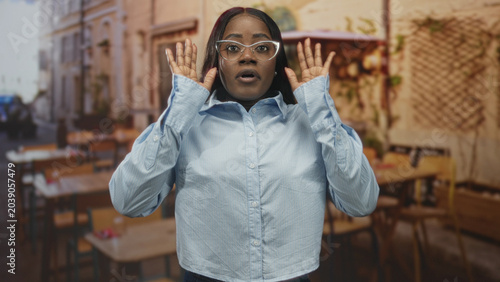 Quadro su tela Woman wearing glasses and blue shirt raises hands palms forward in a street cafe with tables and chairs; surprise caution