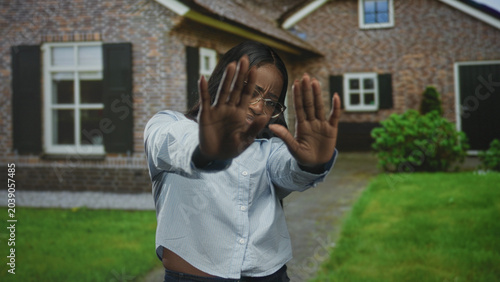 Foto Woman holds up palms to block camera, hands splayed shielding face in front of building with brick facade, window and lawn; privacy