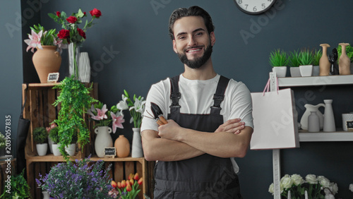 Canvas-taulu Man florist holding pruners with crossed arms among vases and flowers in building shop interior, smiling at camera; friendly confidence