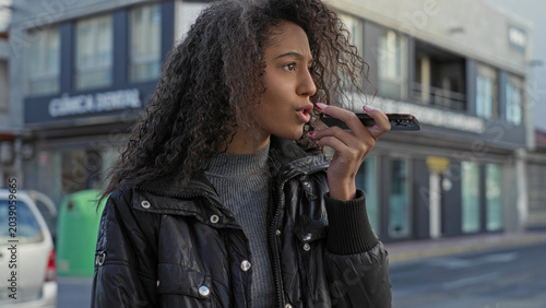 Bild auf Leinwand Teenage woman with curly hair holds smartphone to mouth speaking into speakerphone on street near storefront; concern connection