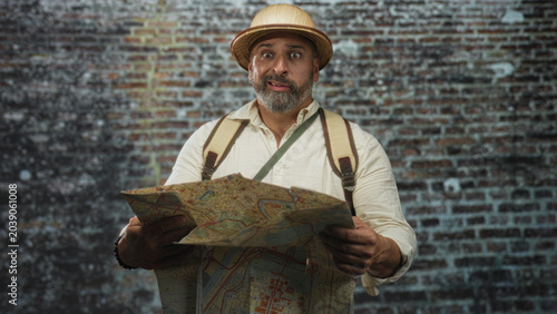 Fotografie Man holding map wearing pith helmet and backpack studying map in a building; confusion travel adventure
