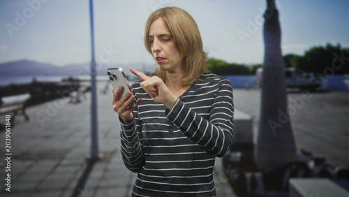 Foto Woman tapping smartphone with finger and frowning while checking messages on a seaside street; frustration technology difficulty