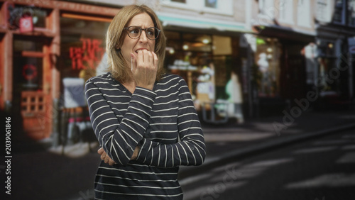 Cuadro en lienzo Woman biting her finger and clutching her other hand, wearing glasses and striped shirt in front of a storefront on a street; anxiety hesitation