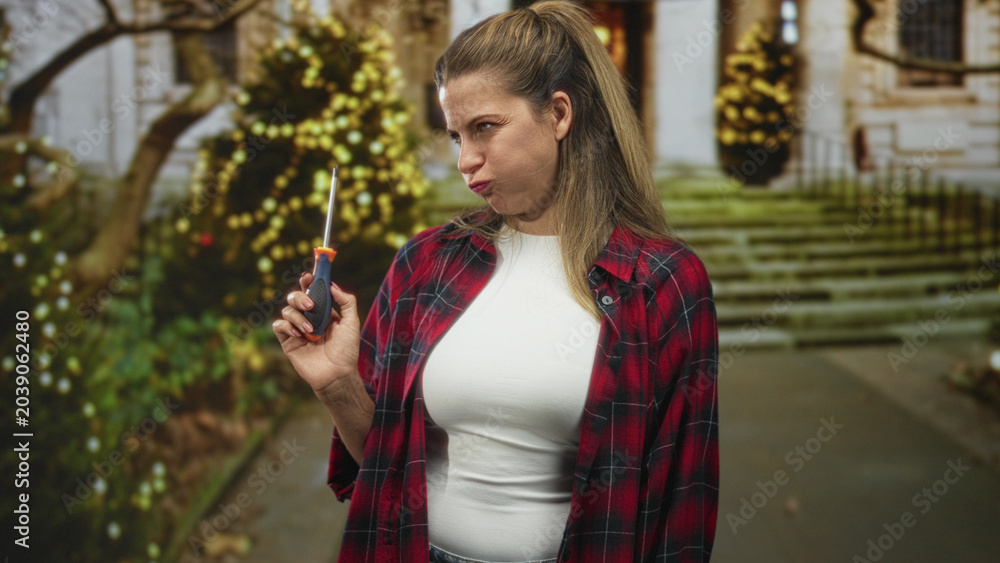Fototapeta premium Woman holds screwdriver in right hand, smiling in a white top and red plaid shirt at a building entrance with steps and holiday lights; cheerful confidence.