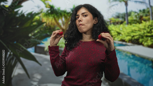 Konstfotografi Woman holding strawberries near face at a hotel pool with puckered lips and closed eyes; playful curiosity