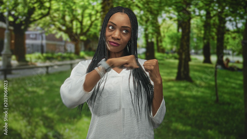 Foto Woman raising fists in forest park wearing white top, bracelets and smartwatch, facing camera with braided hair and assertive pose; confidence resilience