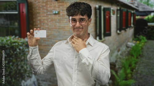 Konstfotografi Young man holding a white chip card near face, hand to throat for choking at building entrance beside brick wall and window outdoors; anxiety