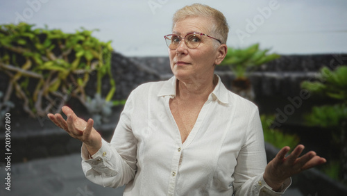 Photos Woman palms up gesture wearing glasses and white shirt near green plants and stone wall in front of building; uncertainty question