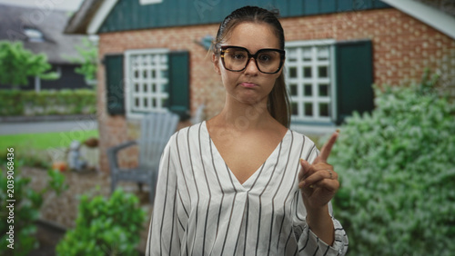 Schilderij op canvas Woman in striped blouse wearing large glasses points finger to the right at a house building entrance outdoors; disapproval judgment