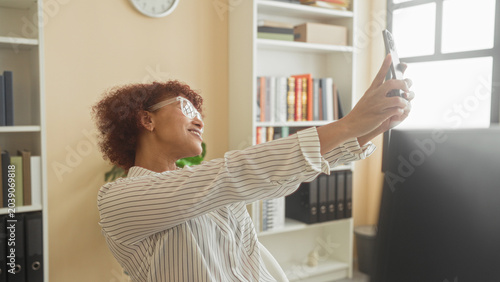 Fotografi Woman holding smartphone smiling and raising arm to take selfie at desk with computer and bookshelves in building; workday joy