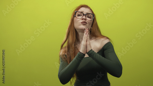 Konstfotografi Redhead woman with hands pressed together in a pleading praying gesture wearing glasses and longsleeve off shoulder top in studio; humble pleading