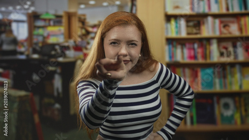 фотографія Redhead woman points finger with pursed lips toward camera among bookstore shelves inside a building; disapproval sternness