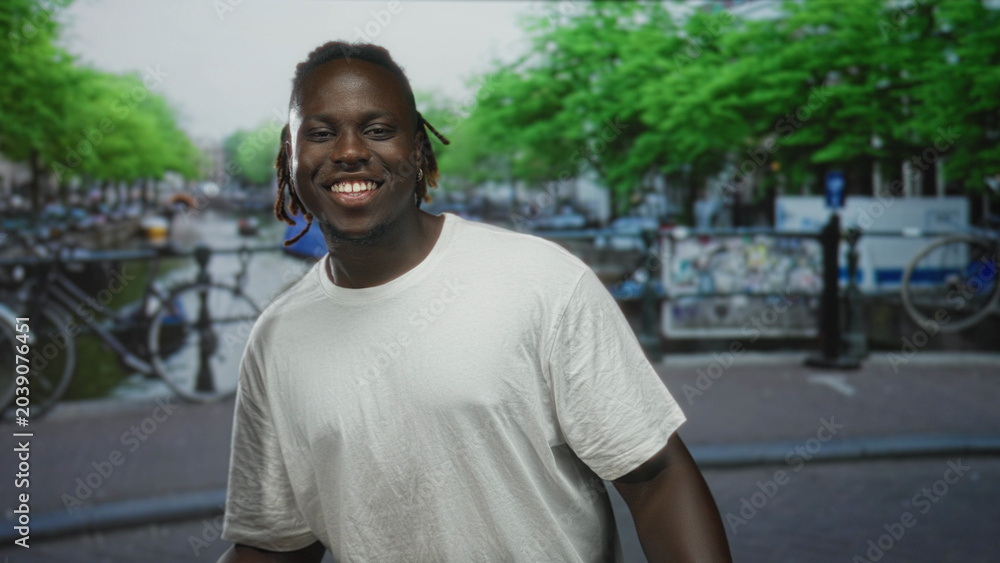 Fototapeta premium Man smiling showing teeth on street beside an amsterdam canal with bicycles and railing; urban summer joy.
