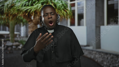 Quadro em tela Man priest yawning with hand to mouth showing clerical collar and cross near building courtyard; tired contemplation