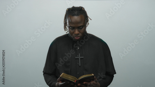 Slika na platnu Young african american priest in black cassock reading a gilded book, holding it with hands in a pale gray studio; faith contemplation devotion