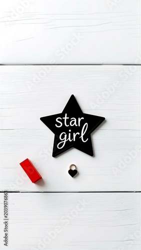 Black star-shaped badge reading star girl with a tiny black heart lock and small red block on white painted wood, minimalist flat-lay accessory conveying girly chic style and ambition