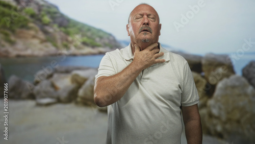 Photos Man holding throat with hand in choking gesture on rocky beach, eyes closed and head tilted back; distress