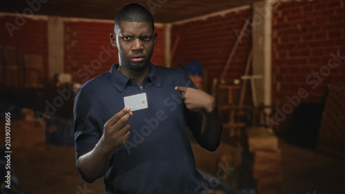 Bild auf Leinwand Young black man holding a creditcard, pointing finger at the card and then to his chest inside a dim brick building storage room; identity doubt