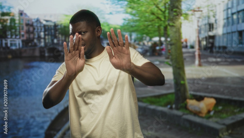 Bild auf Leinwand Man holding palms forward in stop gesture on street canal walkway by water, facing camera with guarded expression; rejection