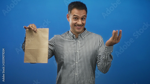 Foto Young hispanic man holds a brown paper bag with right hand and beckons with left hand in studio; playful offer
