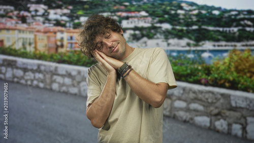 Fotografía Young hispanic man presses hands to cheek in a sleeping gesture on a street beside a stone wall and marina; calm restfulness