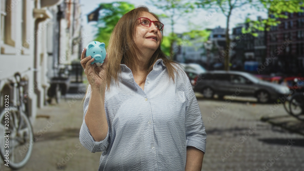 Fototapeta premium Senior hispanic woman wearing glasses holding a blue piggy bank in her hand on a street with parked cars and bicycle; financial planning hopeful.