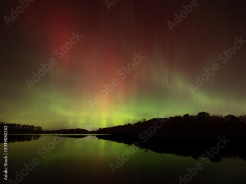 Northern lights of red and green fill the sky and reflect in the waters of a lake below