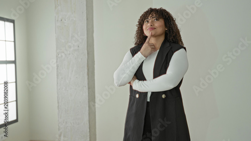 Woman with hand on chin and crossed arm, glancing up while standing by tall window and concrete pillar in empty building interior; thoughtful curiosity.