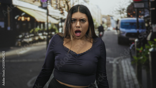 Fotografia Woman shrugs palms up showing cleavage and exposed midriff on a busy street with cafe seating and parked van, mouth open in surprise; confusion doubt