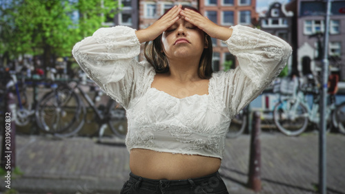 Schilderij op canvas Woman in white lace top showing midriff while bringing hands to forehead on a city street with bicycles and canal; frustration reflection solitude