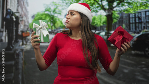 Canvas Print Woman pouting while holding green and red wrapped gifts in hands on street wearing santa hat and red shirt; holiday indecision disappointment