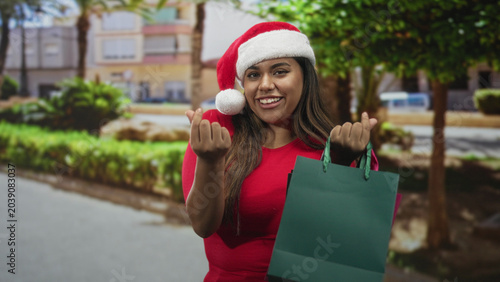 Tableau sur toile Woman in santa hat holds shopping bag and beckons with fingers on street, smiling in red top; cheerful holiday