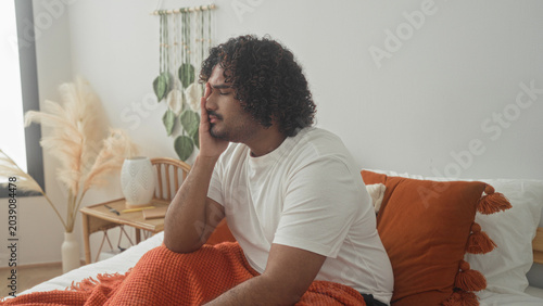 Photos Man sitting on bed covering face with hands, orange blanket and pillow and bedside vase visible inside building; fatigue introspection