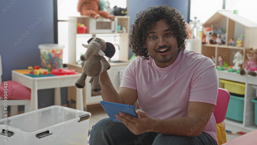 Fototapeta premium Man holds elephant puppet and tablet, points puppet with hand while smiling and seated in building; playful teaching.